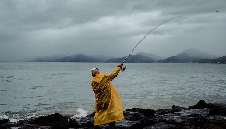 Person in raincoat standing on rocky edge of water casting